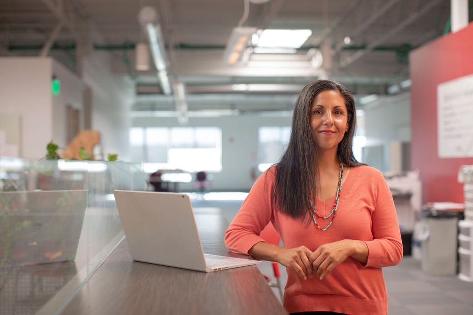 A woman leans against a table next to her open laptop in a large, industrial-looking workspace. She is smiling.